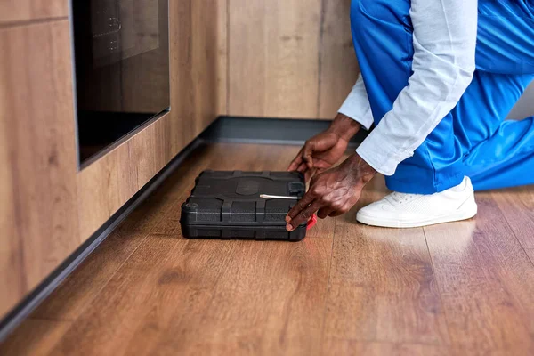 Gekropte weergave van afro zwarte werkman in blauwe overall opening gereedschapskist op keukenvloer — Stockfoto