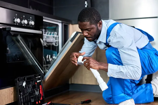 Серьезный молодой африканский рабочий чинит посудомоечную машину, меняет Сифон — стоковое фото