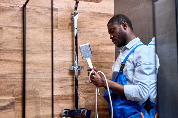 Zijaanzicht op zwarte reparateur klusjesman in blauwe overall met douche handvat in handen — Stockfoto