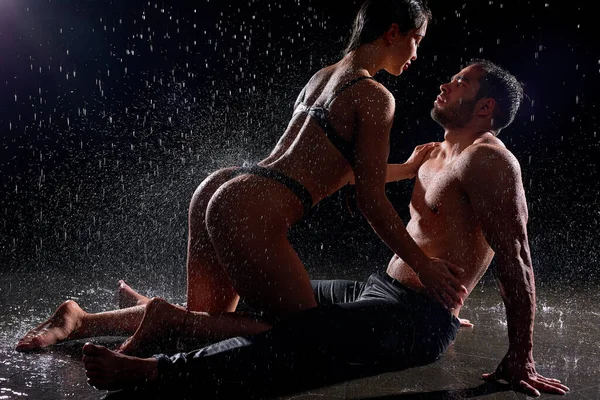 Flirtande par sitter tätt ihop i regn i mörk ateljé, våta och sensuella Stockfoto