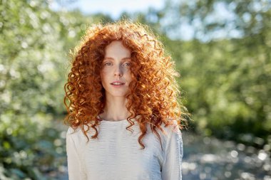 Kıvırcık saçlı romantik kadın kameraya poz veriyor, beyaz elbise giyiyor.