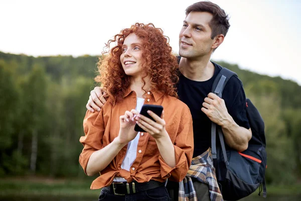 Sonriente pareja de viajeros de pie junto al río utilizando el teléfono inteligente, mirando en el mapa en línea — Foto de Stock