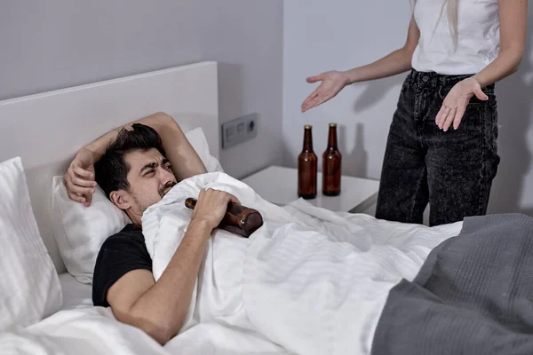 Hombre alcohólico acostado en la cama con alcohol mientras una mujer enojada lo regaña — Foto de Stock