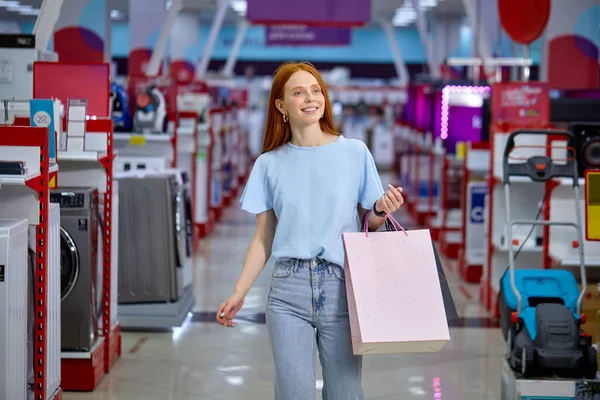 Retrato de mulher andando na loja passando por eletrodomésticos, segurando sacos — Fotografia de Stock