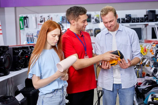 Red-haired female standing with hair dryer in store while husband talking with consultant — Stock Photo, Image
