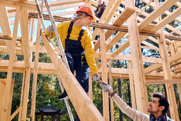 Bebaarde man in uniform helpt vrouw constructeur staan in hoogte, man geven hamer — Stockfoto