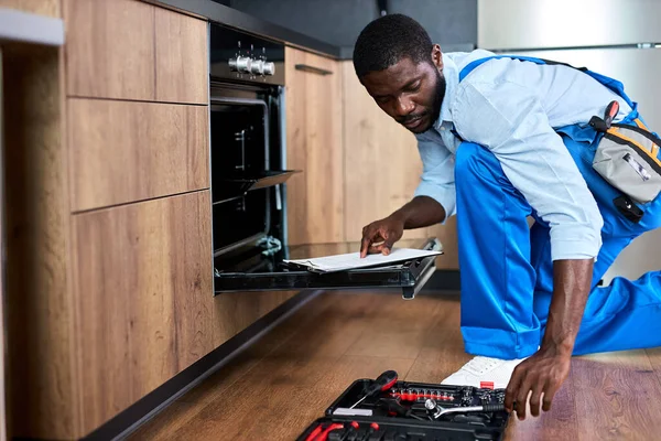 Vertraag niet met repareren. Zijaanzicht afro reparateur onderzoek oven in keuken — Stockfoto