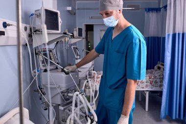 Tıbbi ekipmanların yanında duran erkek doktor, hastanın kalp atışlarını kontrol ediyor.