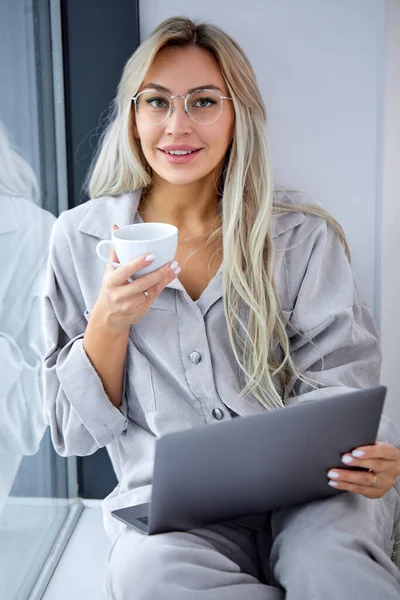 Молодая красивая леди сидит у окна с ноутбуком, использует компьютер дома на подоконнике — стоковое фото