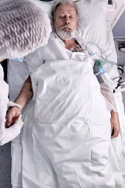 Vista dall'alto sul maschio sdraiato sul letto con tubo endotracheale, medico femminile che controlla l'impulso — Foto Stock
