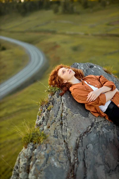 A vörös hajú, göndör nő nagy sziklán fekszik, magas magasságban. A természet és a növényzet látképe — Stock Fotó