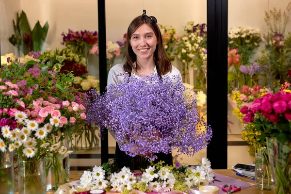 Young woman florist standing in small flowers shop posing, holding many flowers in hands — Stock Photo, Image