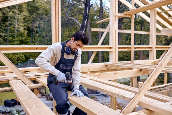 作業服のビルダーは、電気ドリルを使用して、ビームに座って、新しい屋根のツールをインストールします。 — ストック写真