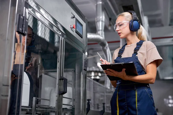 Vrouwelijke fabrieksarbeider houdt kwaliteitscontrole op het werk, neemt notities in folder-tablet — Stockfoto