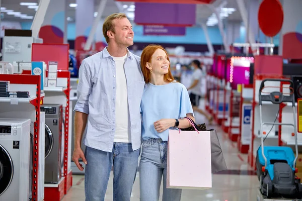 Casal andando na loja passando por eletrodomésticos, segurando sacos de compras — Fotografia de Stock