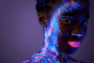 Siyah kadın, neon makyajlı vücut sanatıyla poz veriyor. Kameraya bakıyor, mor renkte izole edilmiş.