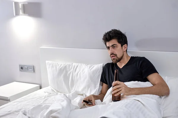 Un hombre en la cama con cerveza viendo la tele. Alcoholismo por aburrimiento durante Covid-19 — Foto de Stock