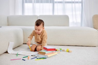 Küçük beyaz çocuk renkli kalemlerle resim çiziyor, yerde oturuyor. Yaratıcılık kavramı.