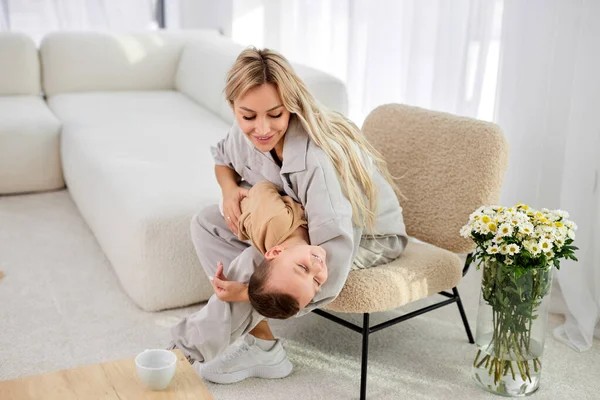 Pozitivní matka se baví se synem v útulném světlém obývacím pokoji, směje se, hraje — Stock fotografie