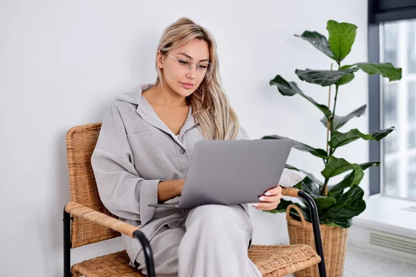 Задумчивая молодая женщина в очках сидит с ноутбуком на диване, смотрит на экран — стоковое фото