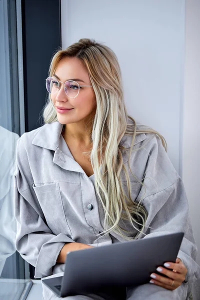 Блондинка в повседневной одежде с помощью компьютера ноутбука и сидя на подоконнике — стоковое фото