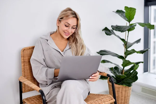 Улыбающаяся женщина в очках, сидящая на стуле с ноутбуком во время работы, общается — стоковое фото