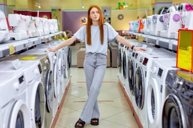 Gayri resmi kıyafetli güzel bayan yeni çamaşır makinelerinin yanında durup, alışveriş yapıyor.