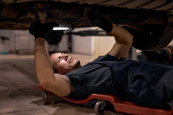 Zkušený mechanik v uniformě pracuje v autoservisu s lampou, na podlaze — Stock fotografie