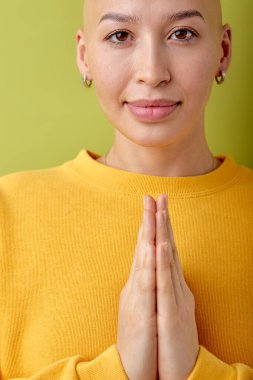 calm and gorgeous young bald woman with smiling face posing at camera, praying. close-up portrait of lady in yellow casual shirt outfit isolated over green studio background, copy space