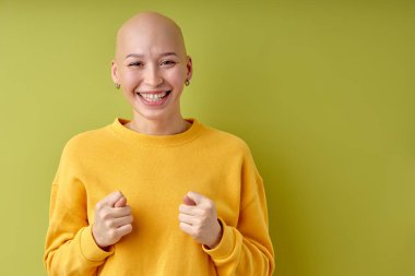 bald happy lady in yellow shirt showing fig gesture, you dont get it anyway. Body language. Refusal fig sign. Young adult woman indoors studio isolated on green wall background, copy space