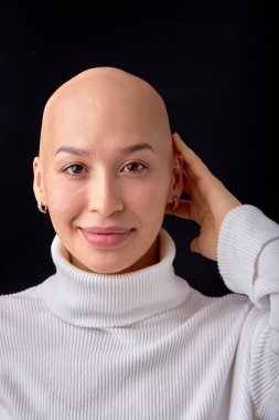 Studio headshot portrait of happy smiling young caucasian woman with shaved head against black studio background. Cancer survivor beauty portrait. Hairless lady in white casual shirt posing