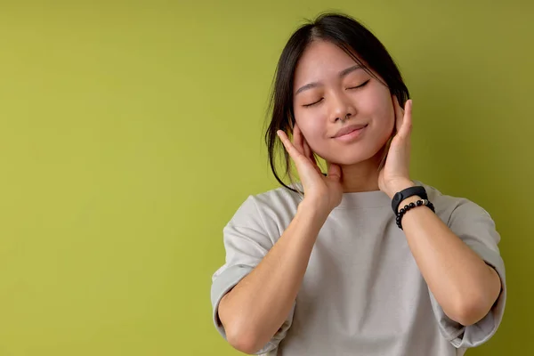 happy chinese woman listening to music in headphones, looking relaxed with eyes closed. portrait of female in casual wear enjoying music, isolated over light green studio background, copy space