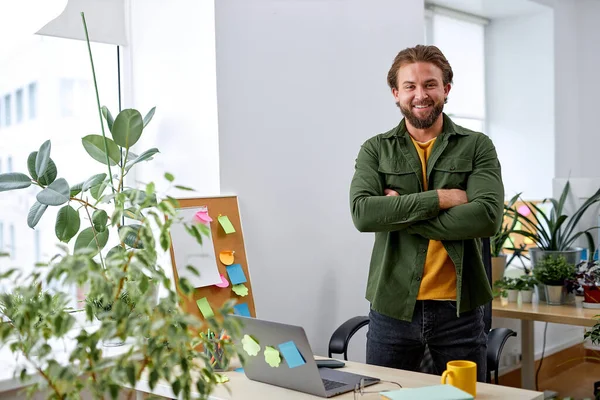 Portrait of excited caucasian manager designer man in office posing at camera, standing behind desk with laptop, many plants in office room. European guy in casual wear, success and business