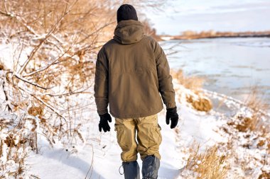 rear view man in warm clothes walking in snowy nature in winter, have rest, enjoy active lifestyle. handsome guy walk along snowy fields. at frozen winter day. alone, outdoors. bushcraft, hike, travel