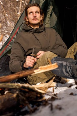 calm handsome man sit cutting wood with knife, next to tent, camping, travel concept. caucasian male in warm clothes having rest, relaxing. alone. bushcraft, adventure, preparing bonfire