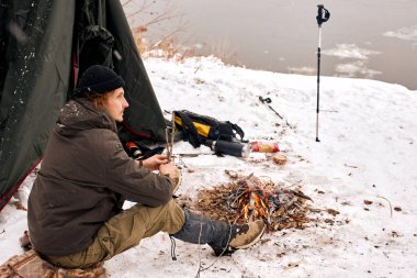 handsome caucasian guy in warm clothes sitting next to tent during camping, alone. young male have rest, relaxing, look at side in contemplation, enjoy the moment in winter forest, nature
