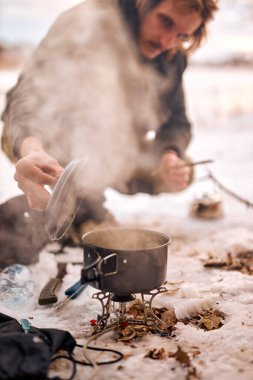 young caucasian guy preparing food in winter forest. Bushcraft, hike, camping, travel, adventure and people concept. smoke from prepared cooked meal on small saucepan. in frozen