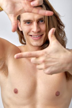 naked fit male with long hair posing at camera, half-naked man looks at camera, making shape of square like a photographer, smiling happily. isolated white studio background. close-up portrait