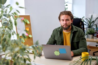 Portrait of attractive young businessman using laptop computer at office desk. looking at camera. caucasian european bearded guy in casual wear outfit is concentrated on work thinking
