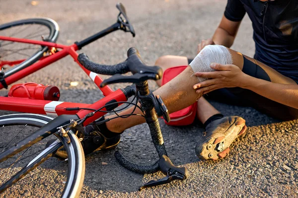 Accident man in sports concept. biker fall from bike and wrapping leg in bandage. cropped injured young male bycyclist in black sportswear has an accident on bike, red bike on road next to him