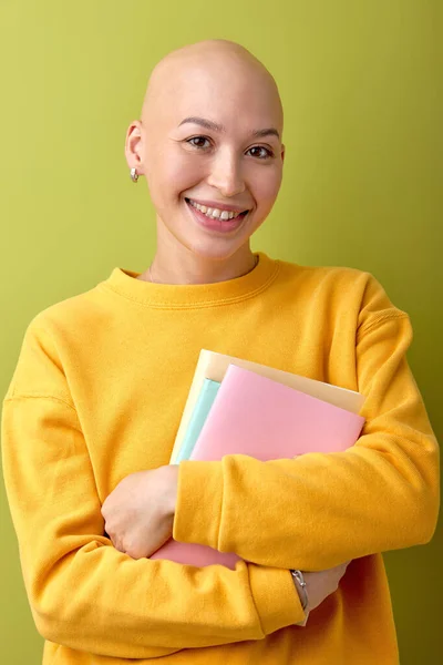 Excited female with bald head holding books in hands and smiling, enjoying education, studying. Hairless lady in casual yellow shirt posing at camera, university concept. people lifestyle