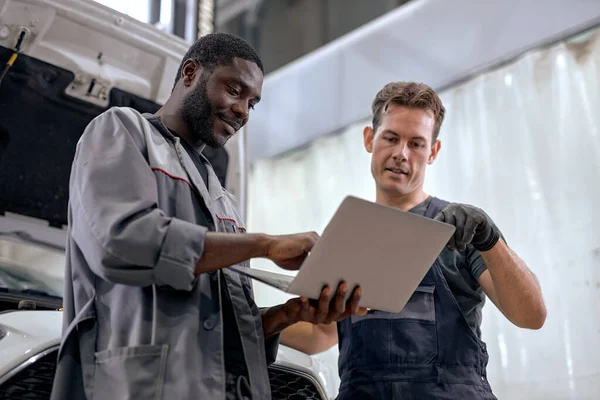 team of diverse mechanics in uniform examining car, using laptop while working in auto service. black and caucasian Auto mechanics checking car,service via online insurance system at garage