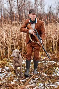 caucasian hunter male holding gun and walking in forest. Rifle Hunter In rural area. Autunm hunting. hunting with dog. A hunter with dog and hunting gun, ready to shoot, in brown stylish suit