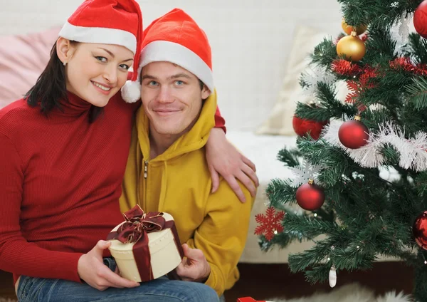 Счастливая молодая пара с подарком сидя рядом с елкой — стоковое фото