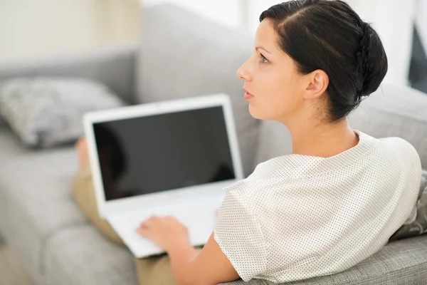 Thoughtful young woman laying on couch and using laptop — Stock Photo, Image