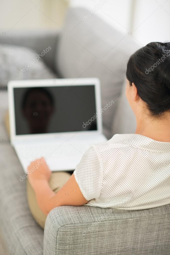Young woman laying on couch and working on laptop. Rear view