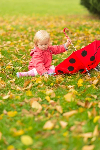 Baby with red umbrella collecting fallen leaves — Stock Photo, Image