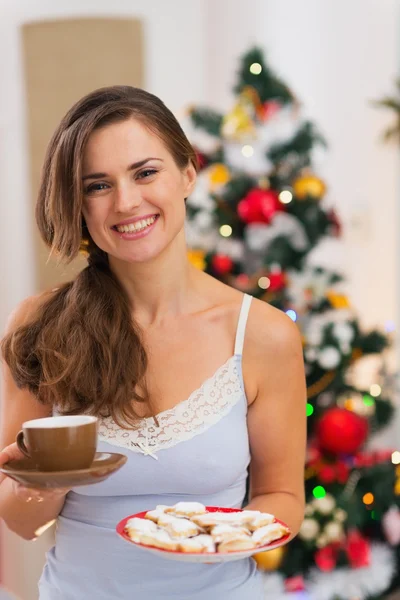 Happy woman in pajamas holding hot beverage and cookies in front — Stock Photo, Image