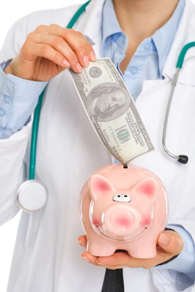 Medical doctor putting 100 dollars note into piggy bank — Stock Photo, Image