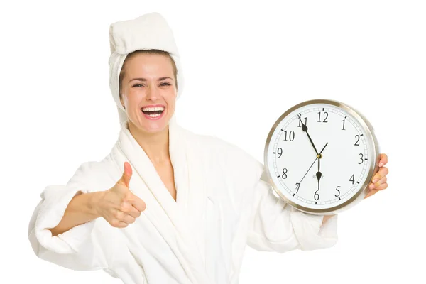 Young woman in bathrobe showing clock and thumbs up Stock Photo